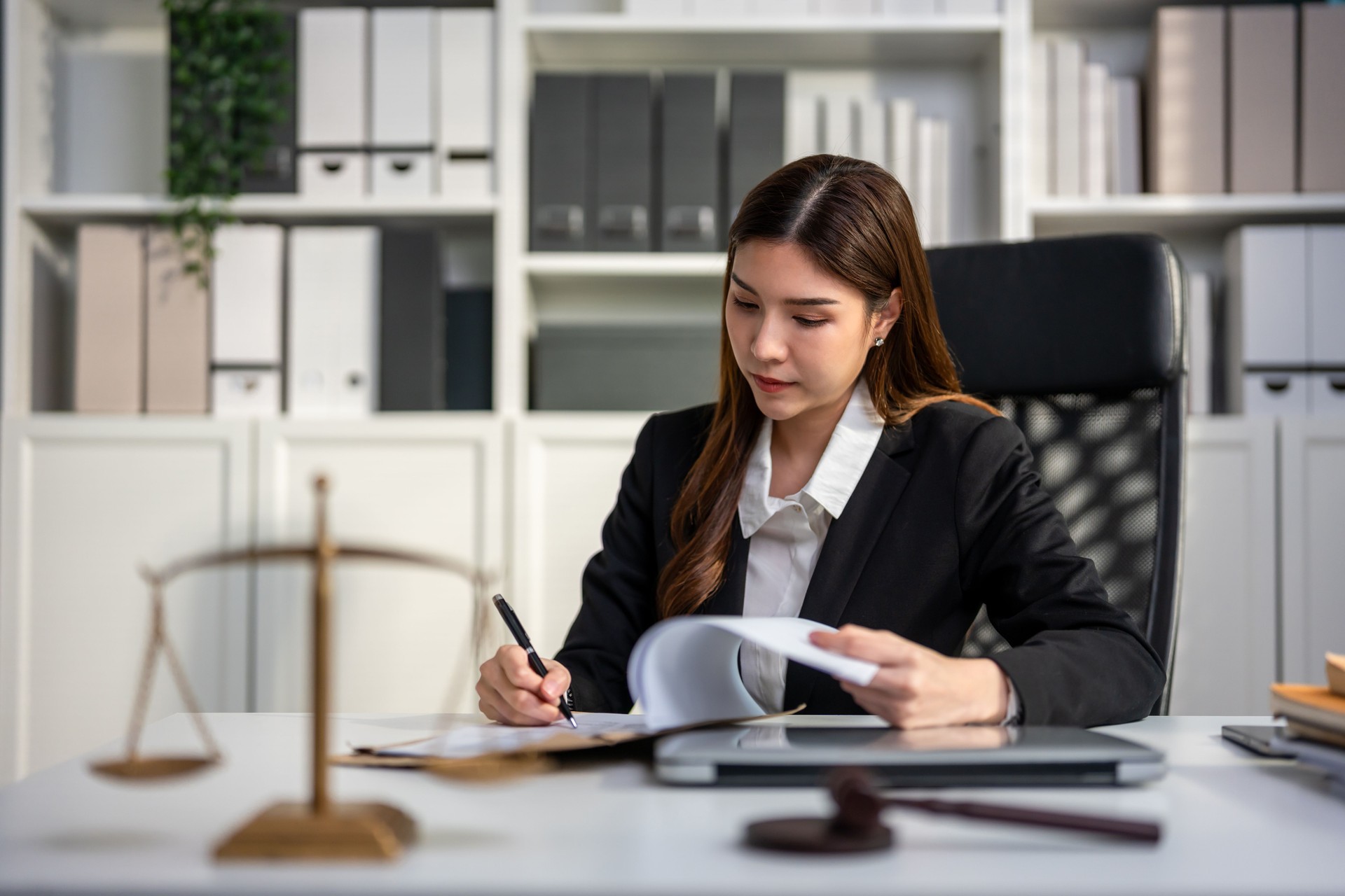 Asian lawyer woman work with hammer and scales of justice in courtroom. Attractive judge attorney looks at paper and document while sitting on table in office. Law and Legal business services concept.